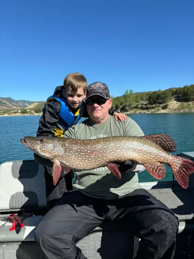 Man and boy on boat hold large fish; blue water, sky; mountains in background.