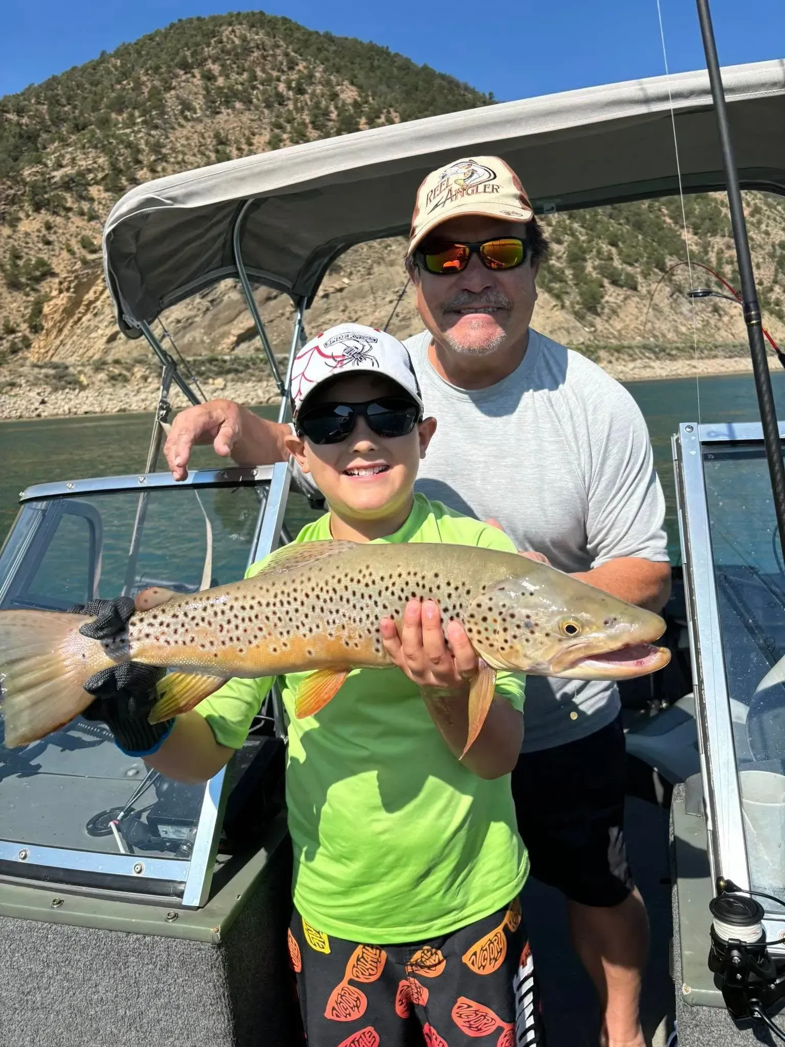 Boy holding large brown trout with man on boat; lake and mountains in background.