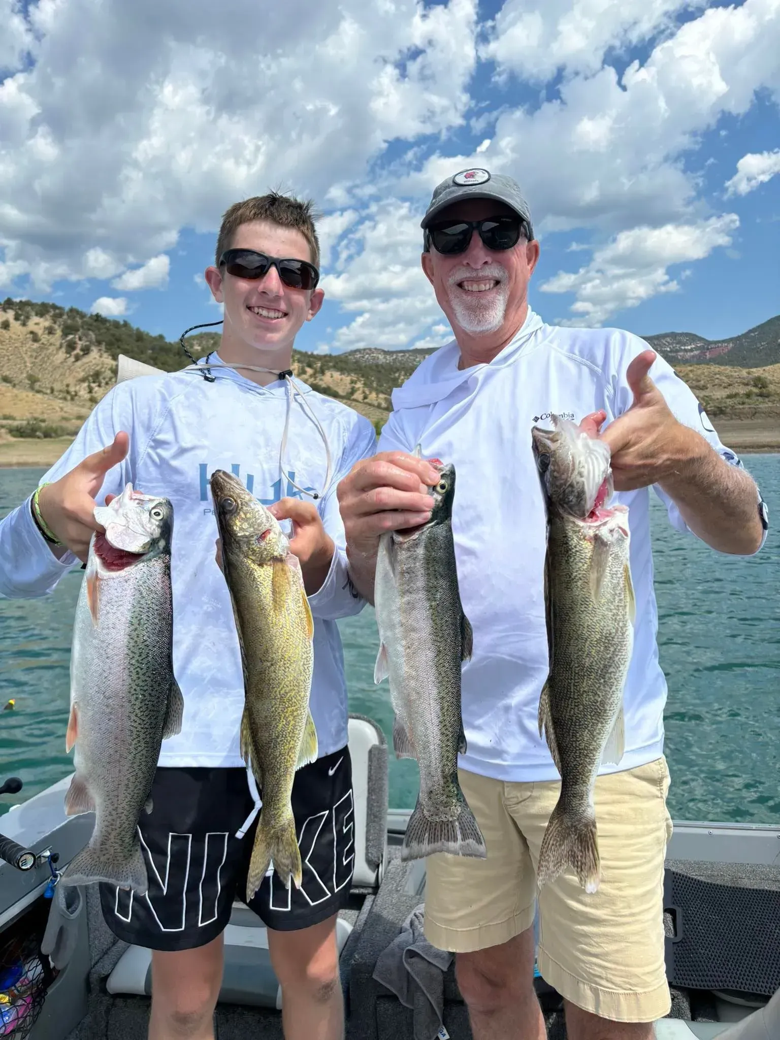 Two men on a boat show off their caught fish. One gives a thumbs-up. Blue water, sunny sky.