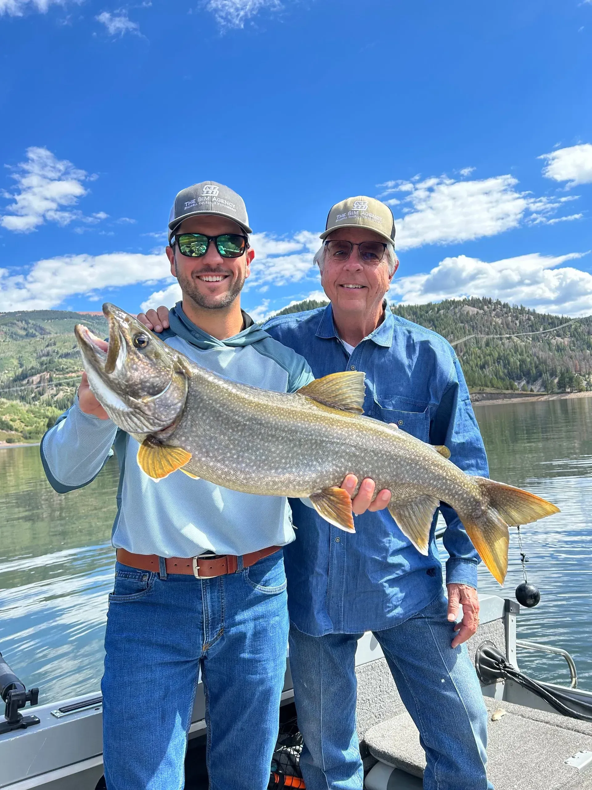 Two men on a boat hold a large fish. Sunny day, lake background, both wearing hats, smiles.