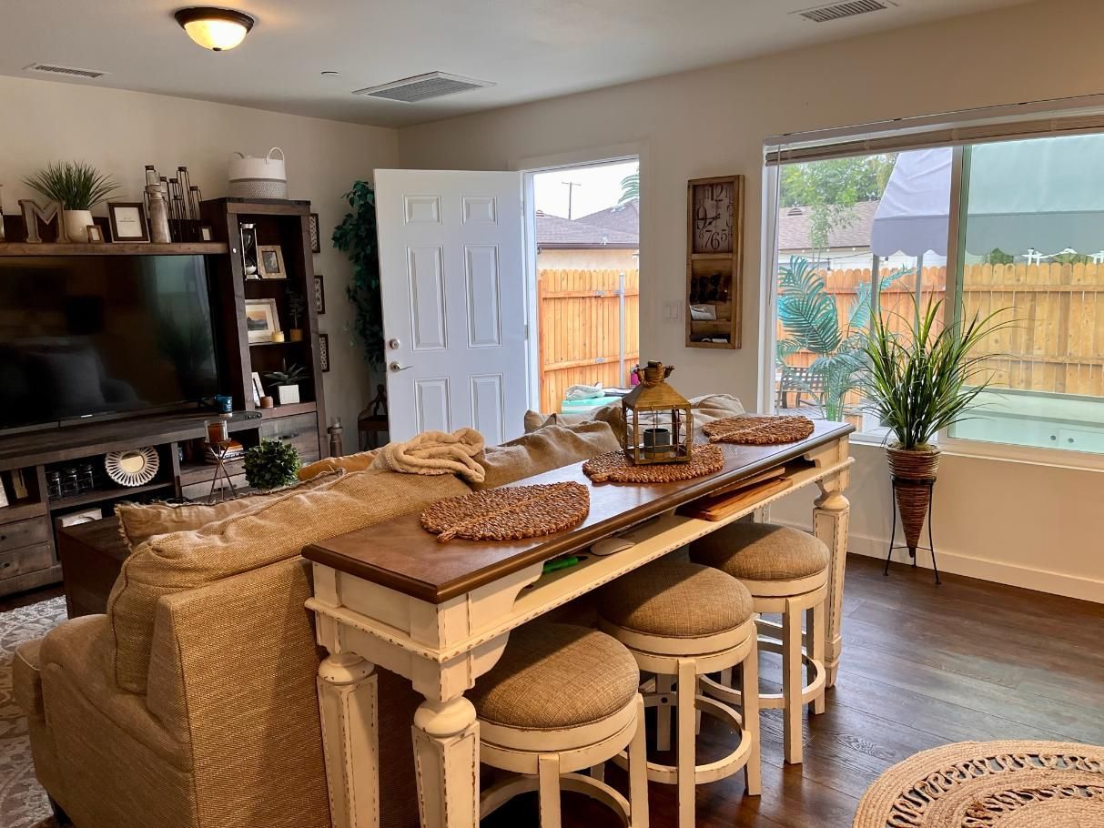 Cozy dining area with a long wooden table, stools, TV, and bright windows overlooking a fenced yard