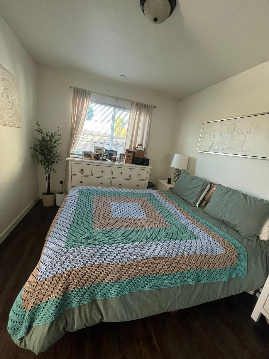 Bedroom with a neatly made bed, teal patterned quilt, window, dresser, and potted plant.