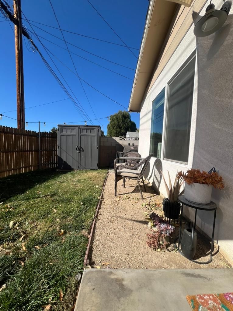 Narrow backyard walkway beside a house, with a gravel path, patio table, shed, fence, and blue sky.