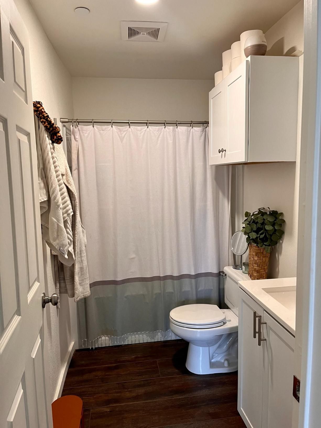 Narrow bathroom with white cabinets, toilet, shower curtain, and dark wood floor