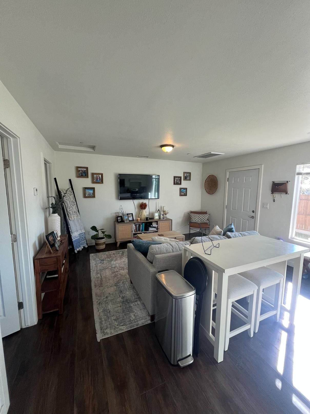 Bright living room with gray sofas, TV on wall, white dining table, and sunlight through blinds