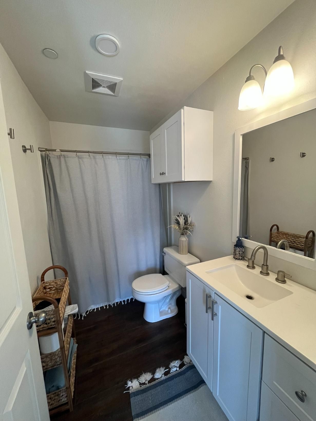 Narrow bathroom with white vanity, toilet, shower curtain, wicker baskets, and twin wall lights.