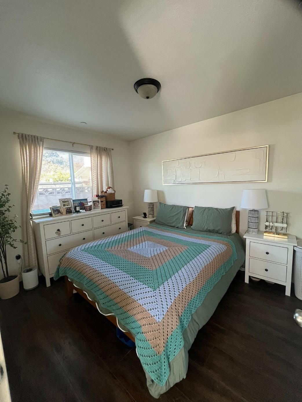Bright bedroom with teal quilted bed, white nightstands, and a window with curtains.