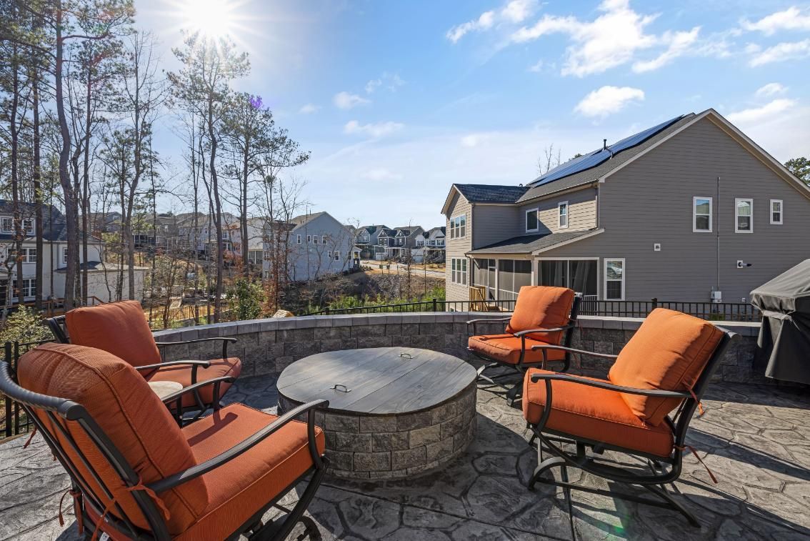 Patio with orange cushioned chairs around a round stone fire pit, overlooking houses and trees under a blue sky