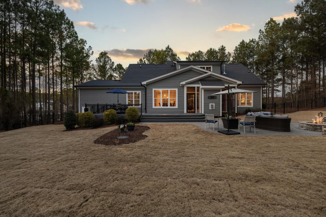 Gray house with lit windows, front porch, and landscaped yard at dusk surrounded by tall trees