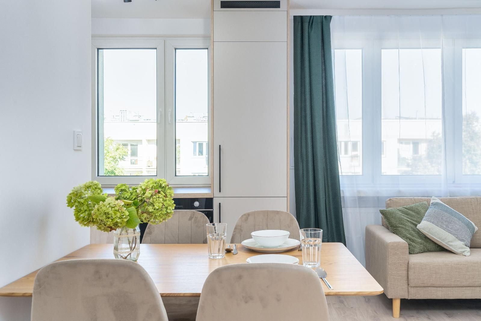 Bright dining and living room with a wooden table, flowers, sofa, and large windows with curtains