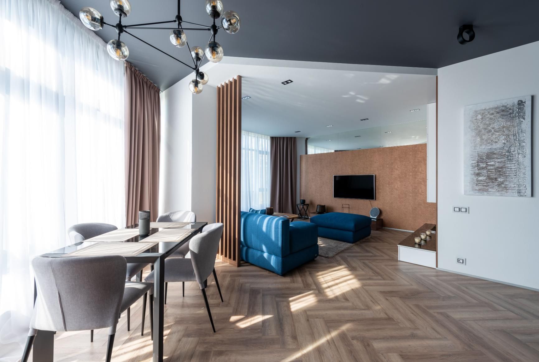 Bright modern living room with blue sectional, dining table, wall-mounted TV, and large windows.
