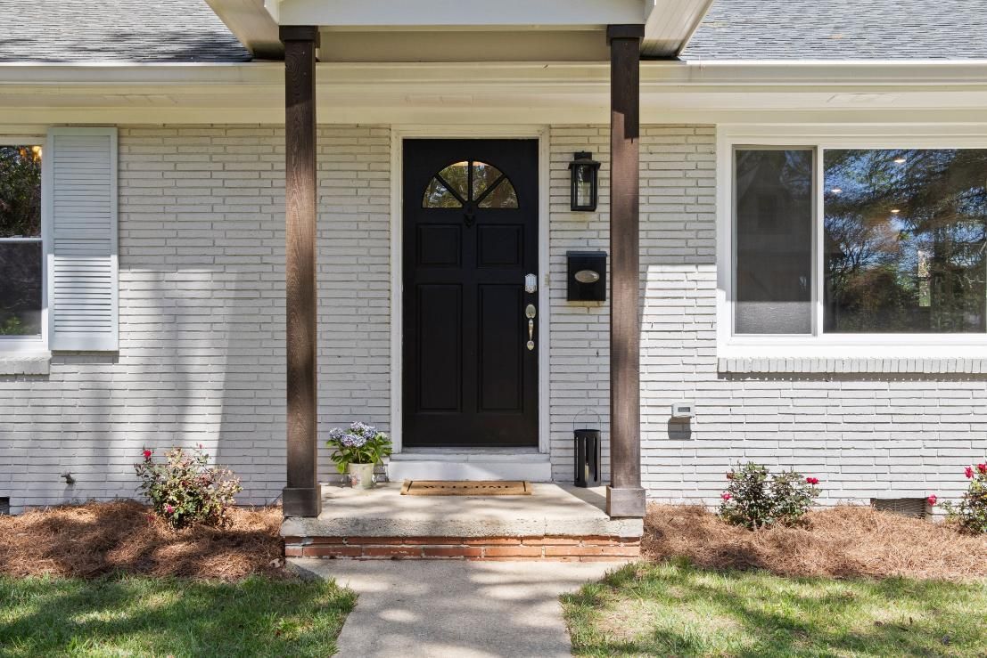 Front porch with black door, white siding, and a small window, bordered by grass and shrubs.