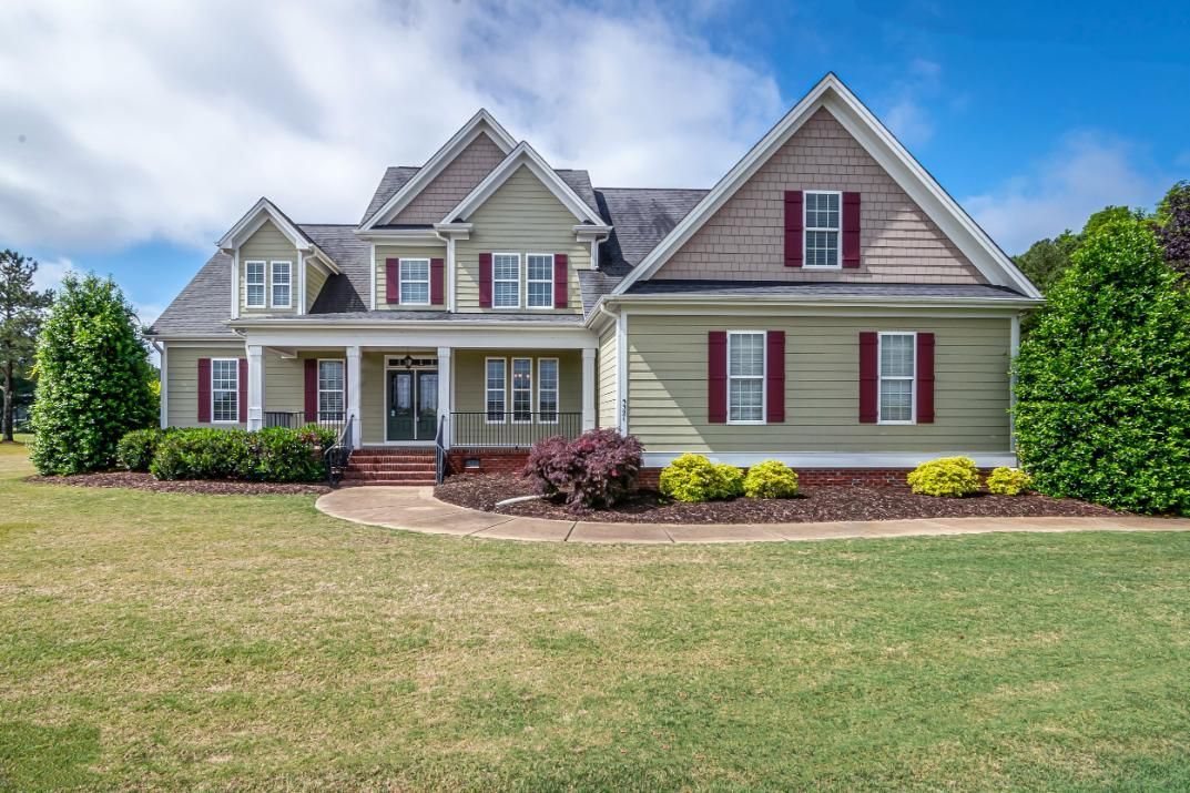 Large beige two-story suburban house with red shutters, white trim, and a broad green lawn under a cloudy sky