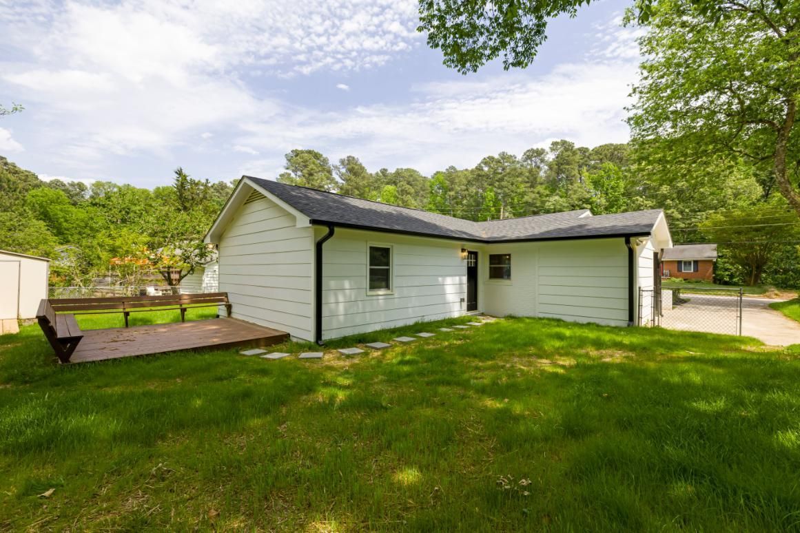 White house with a large green lawn and wooden deck, surrounded by trees under a bright sky