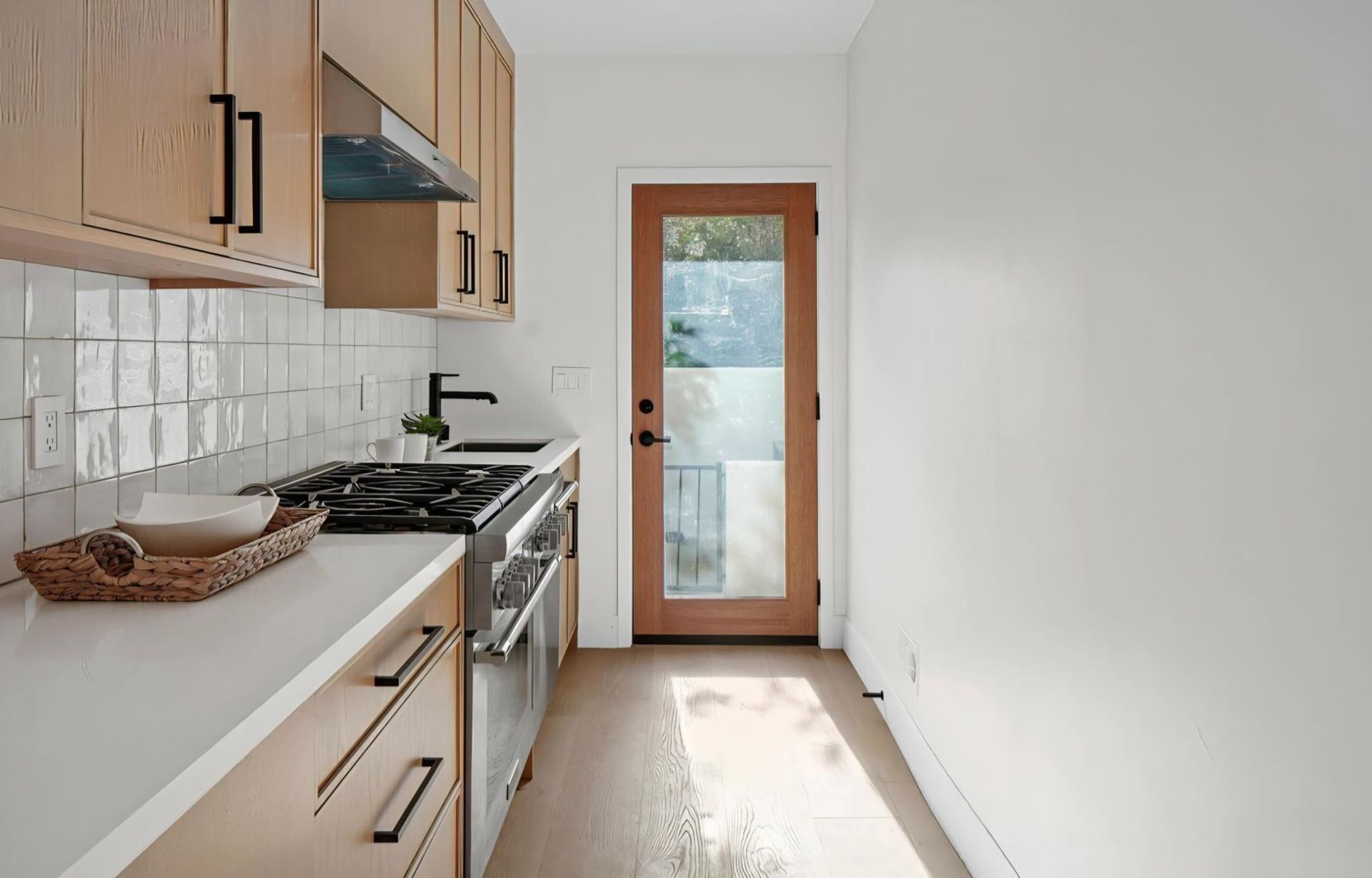 Narrow kitchen with light wood cabinets, white countertops, gas stove, and a door to the outside.