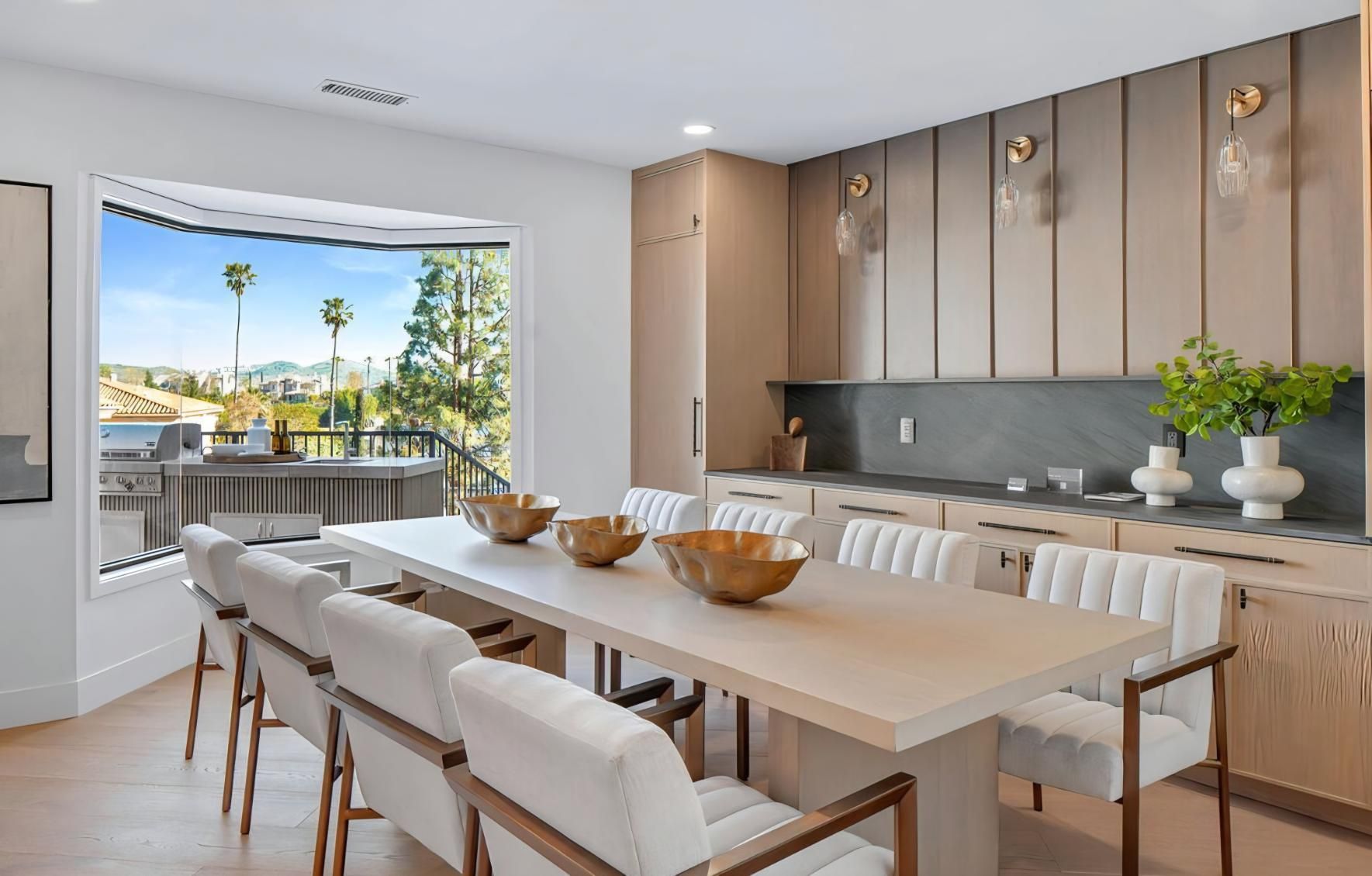 Bright modern dining room with a long table, beige chairs, wood cabinets, and an open view to a balcony.