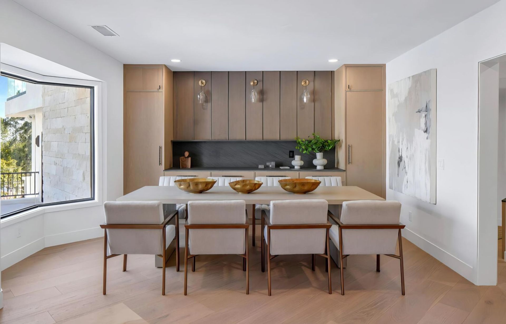 Modern dining room with a long table, beige chairs, wood cabinets, and a large window.