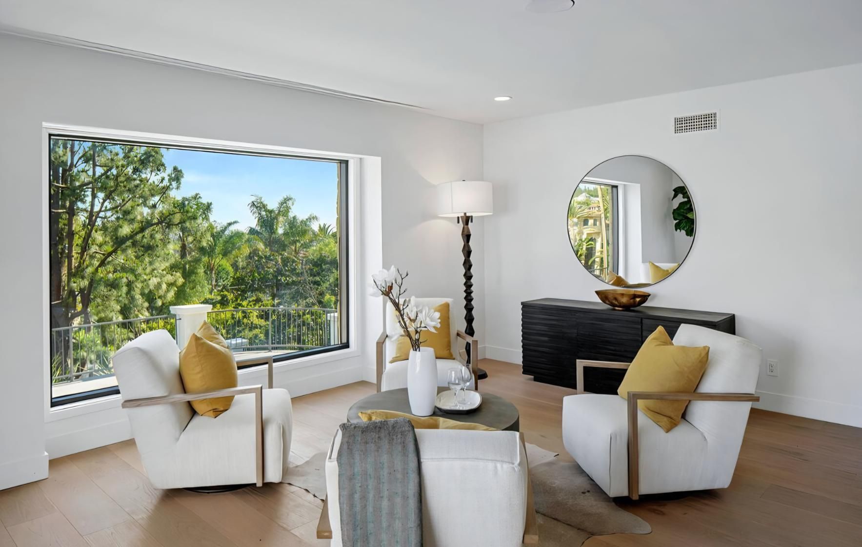Bright living room with white chairs, yellow pillows, a round mirror, and a view of lush greenery outside.