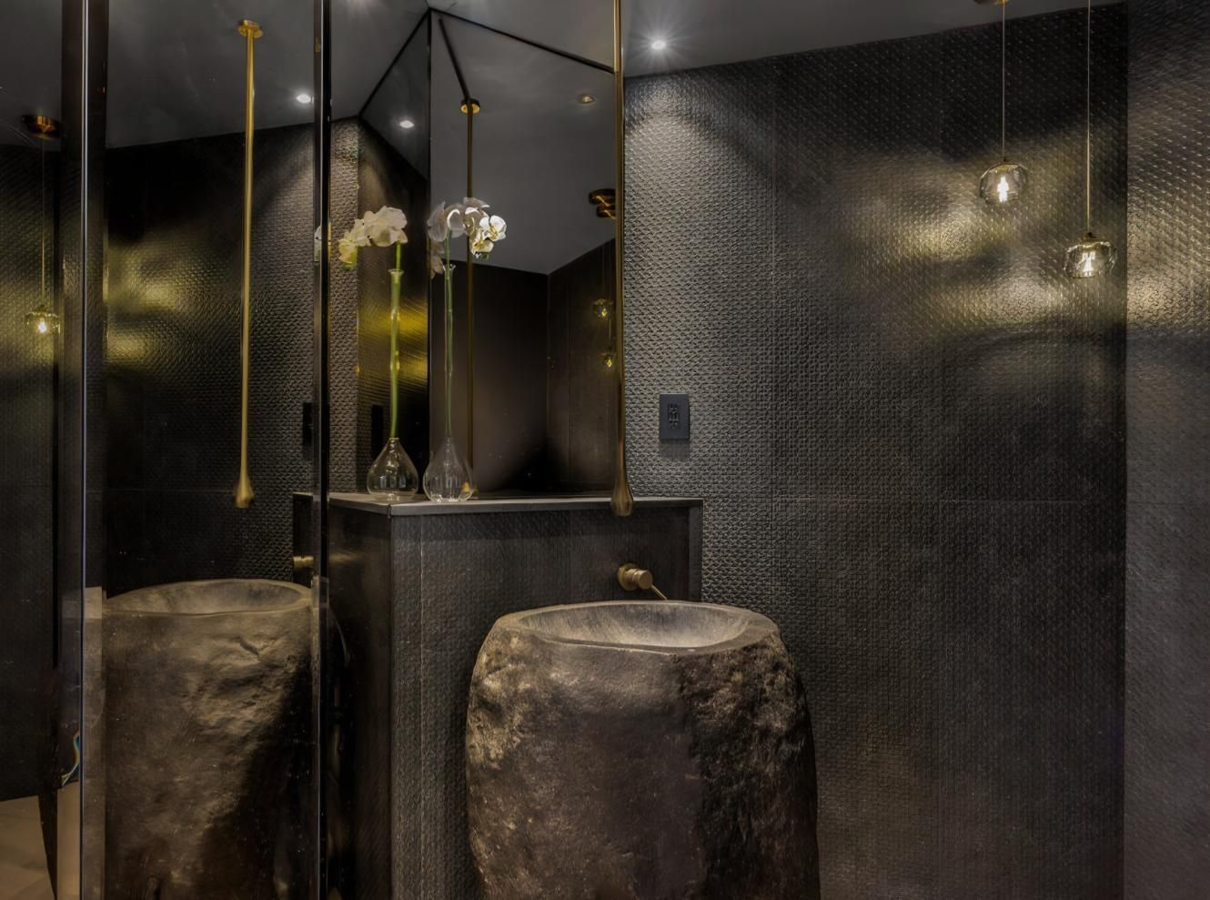 Dark, modern spa room with stone stools, black tiled walls, and hanging gold light fixtures