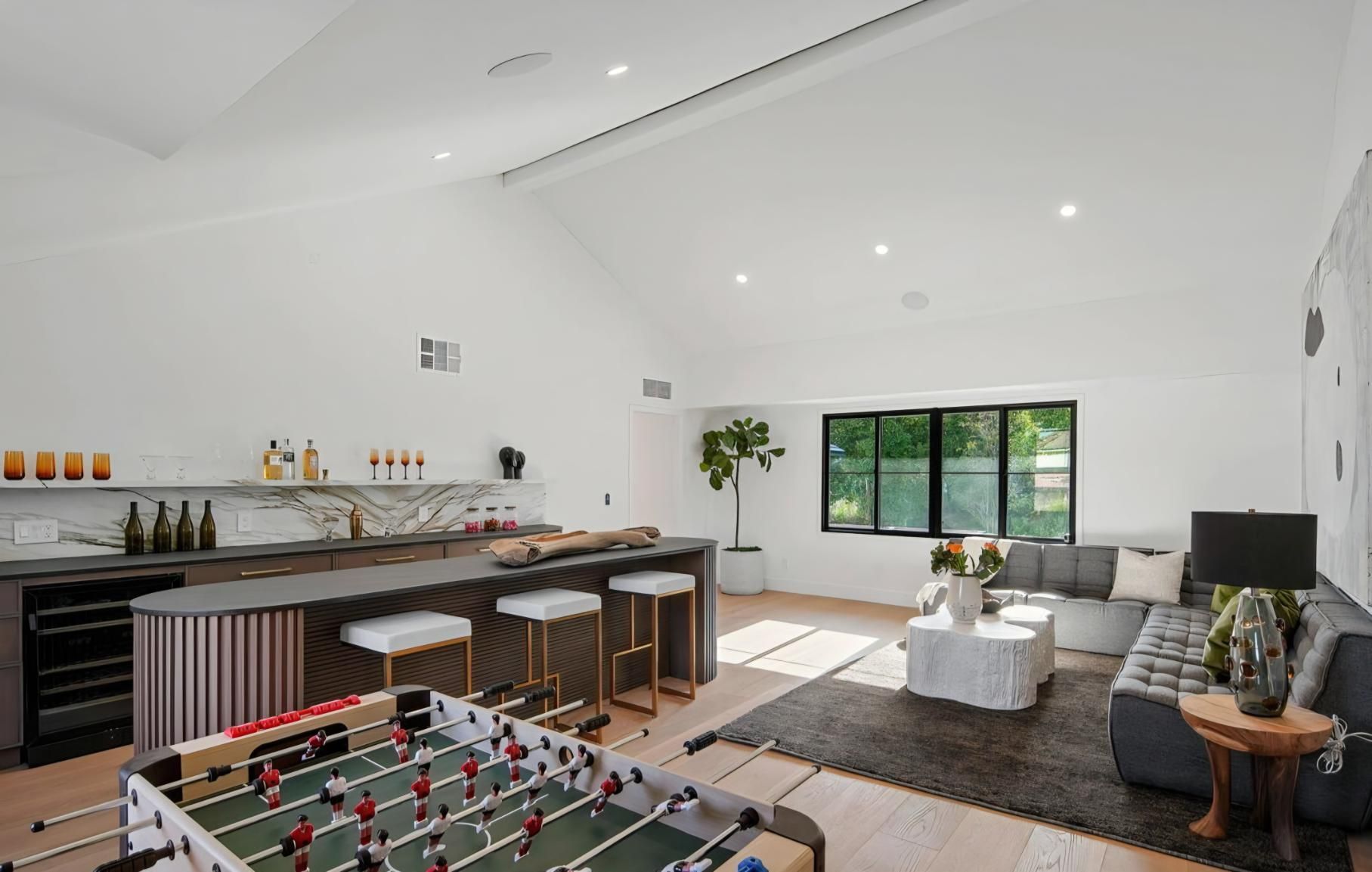 Bright open living room with kitchen, foosball table, gray sofa, and large window.