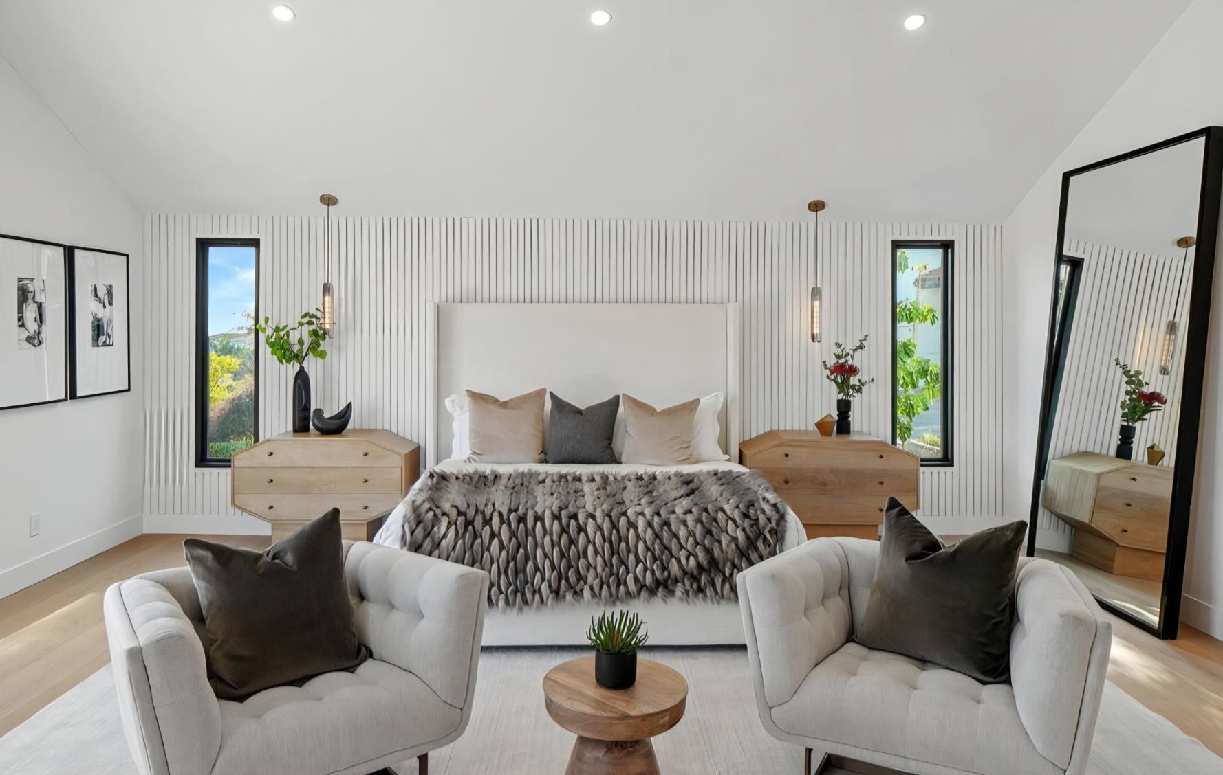 Bright modern bedroom with white walls, twin mirrors, upholstered chairs, and a large bed with patterned bedding.