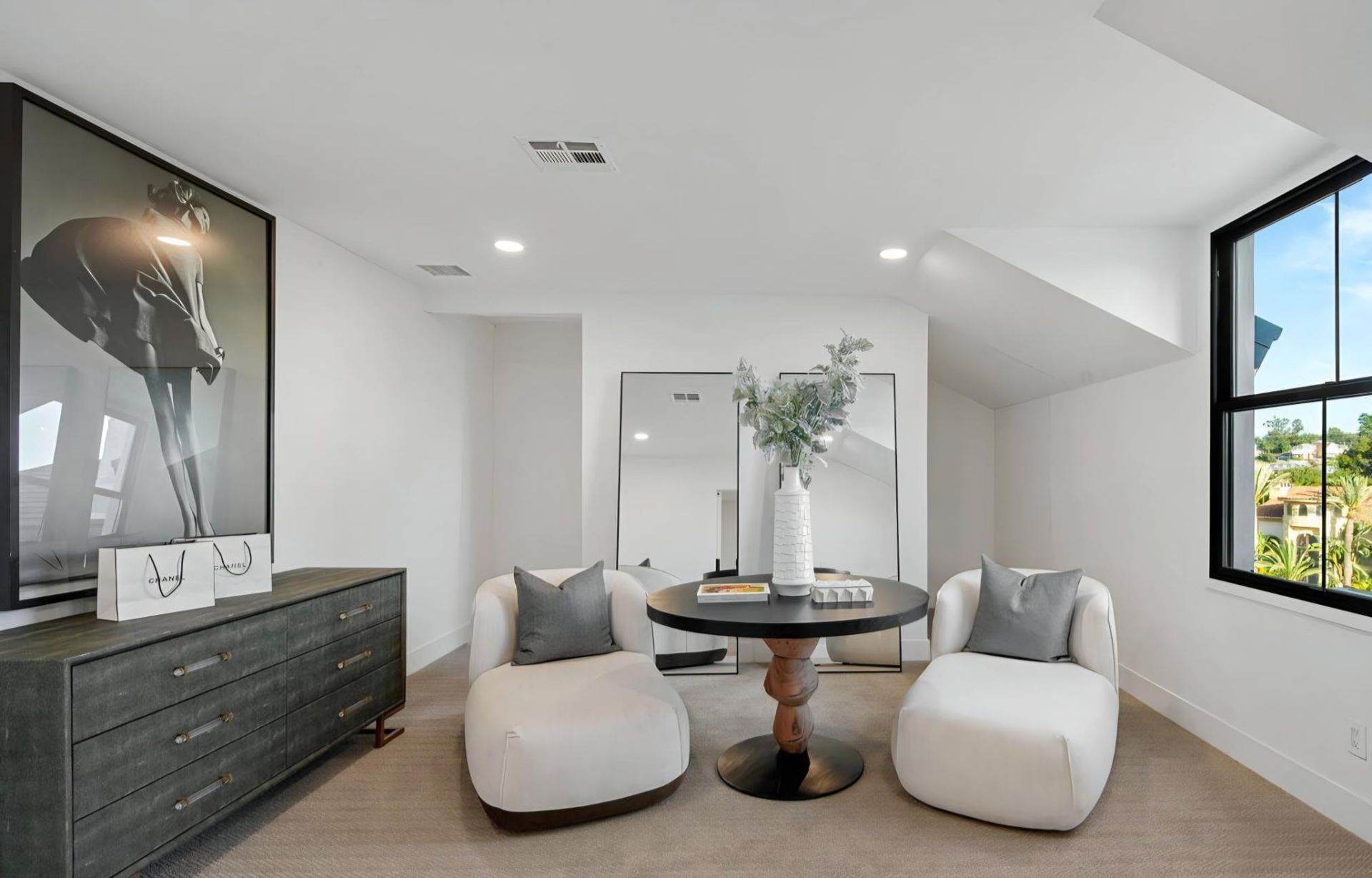Bright modern sitting room with two white chairs, black dresser, round table, and large window