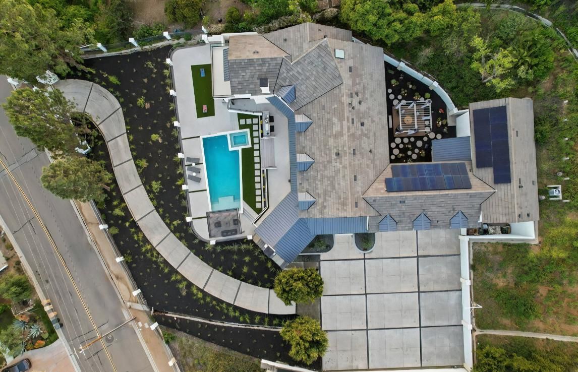 Aerial view of a large house with a blue pool, driveway, solar panels, and surrounding trees.