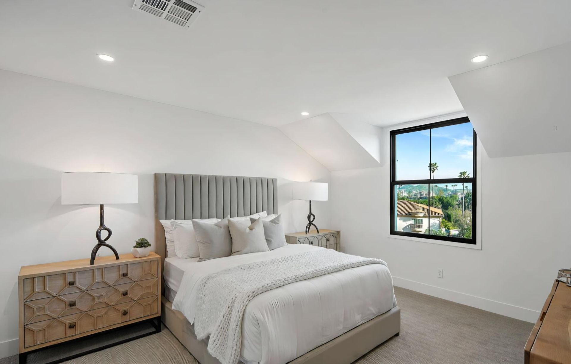 Bright bedroom with a bed, two lamps, a wooden dresser, and a window under a sloped ceiling.
