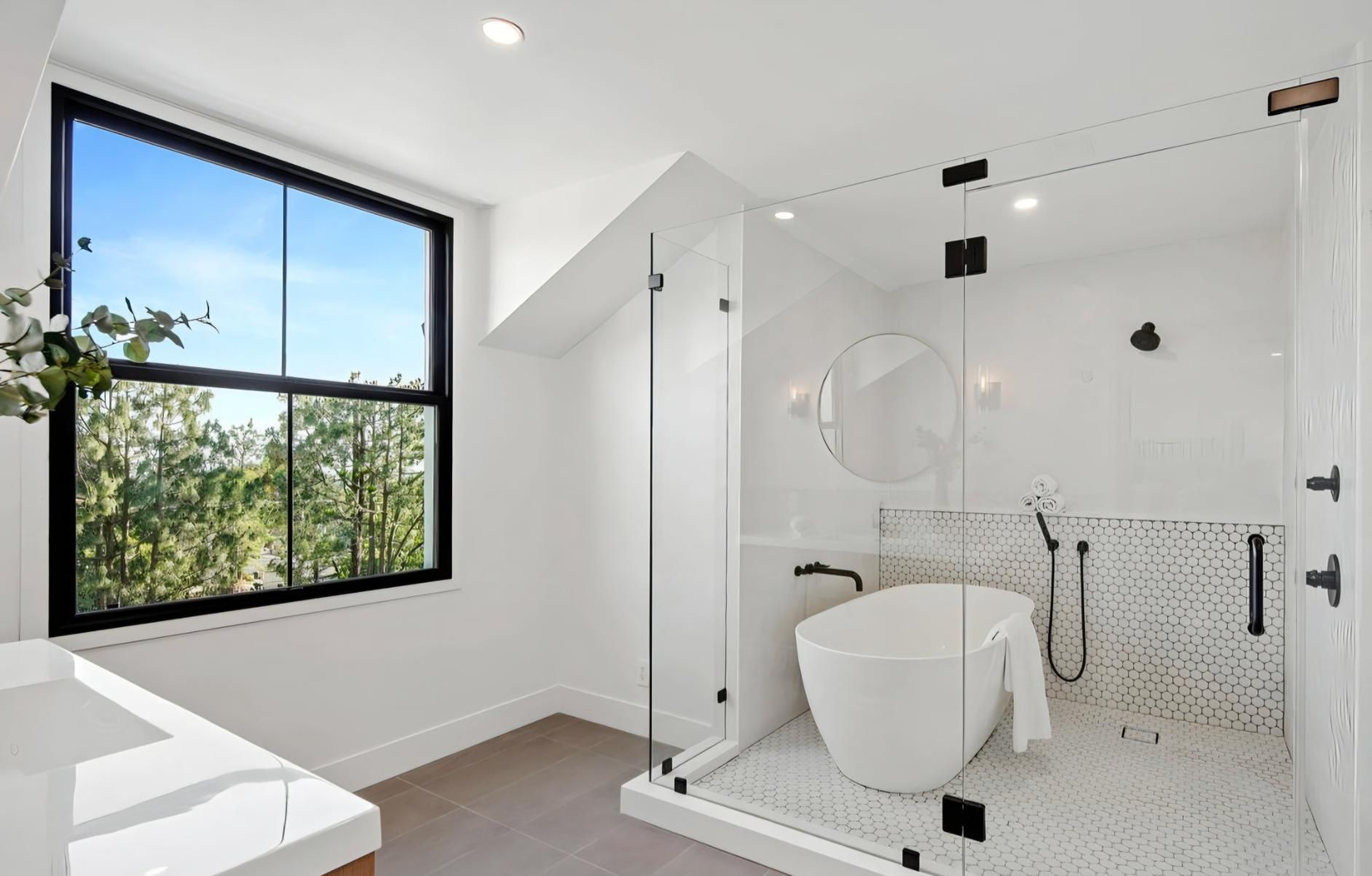 Modern white bathroom with glass shower, freestanding tub, and large window with trees outside