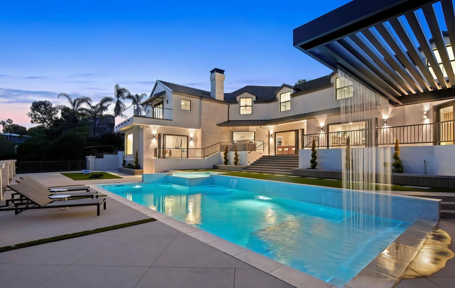 Luxurious modern house with a lit backyard pool at dusk, lounge chairs and waterfall feature