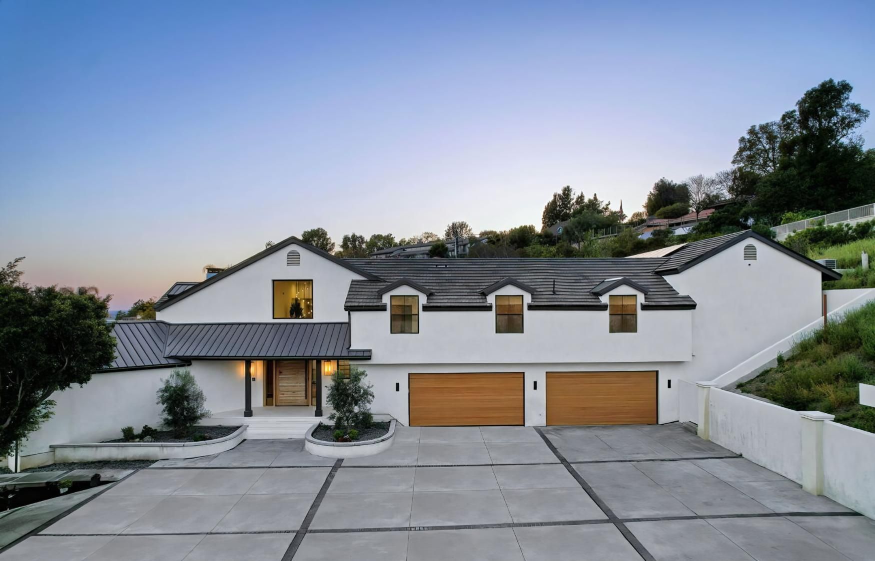 Modern white hillside house with gray roof, warm lights, and a wide concrete driveway at dusk