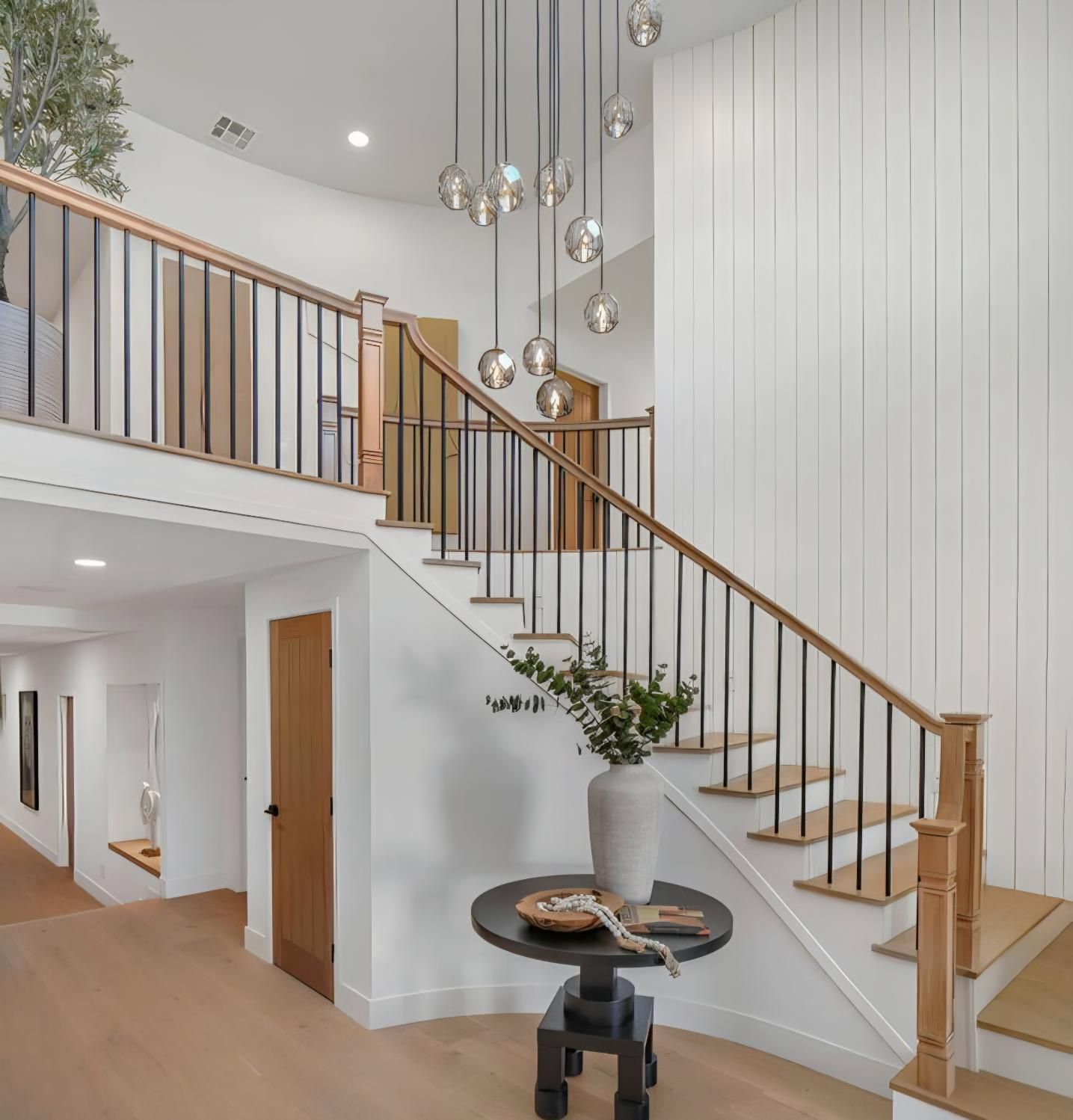 Bright foyer with wood staircase, white paneled wall, hanging glass pendants, and a small round table with a vase.