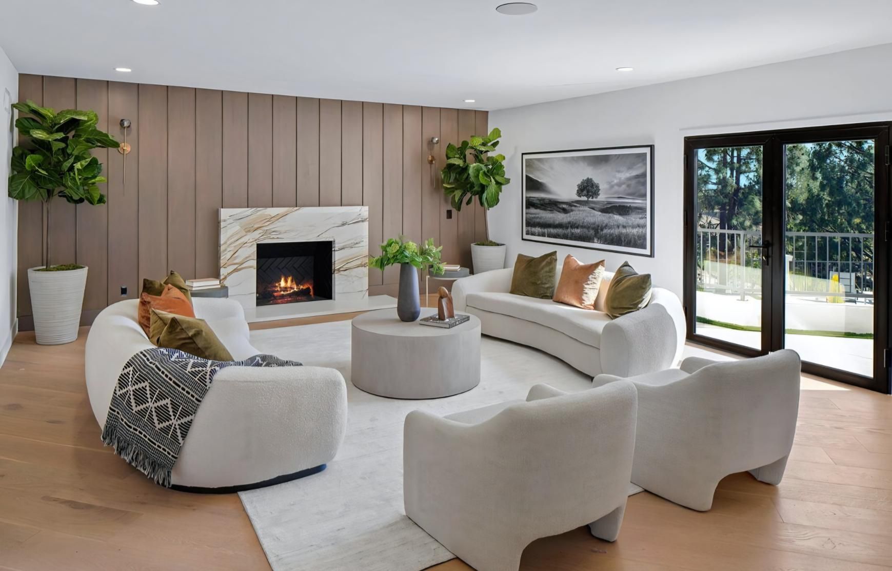 Modern living room with white sofas, fireplace, plants, and large glass doors to a sunny patio