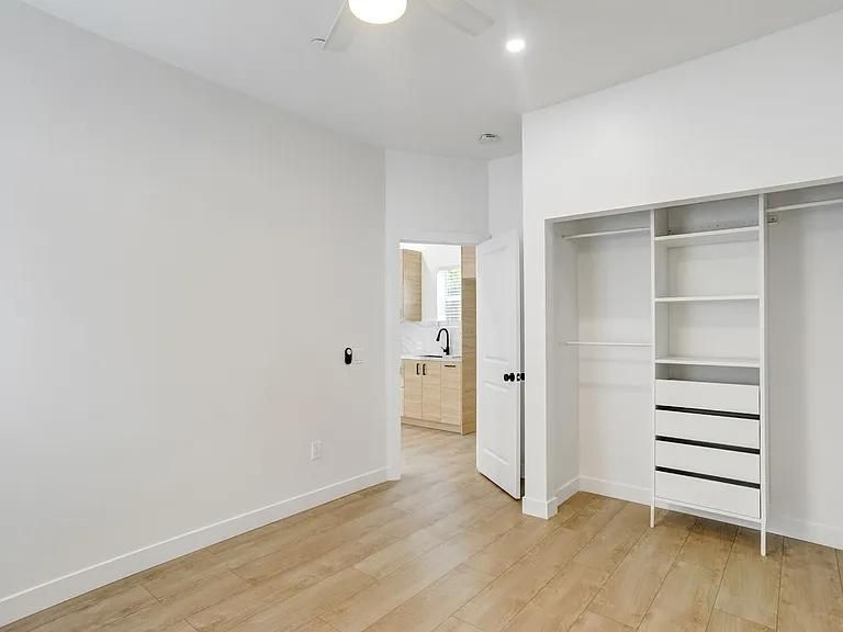 Bright empty white bedroom with wood floor, open closet, and doorway to bathroom.
