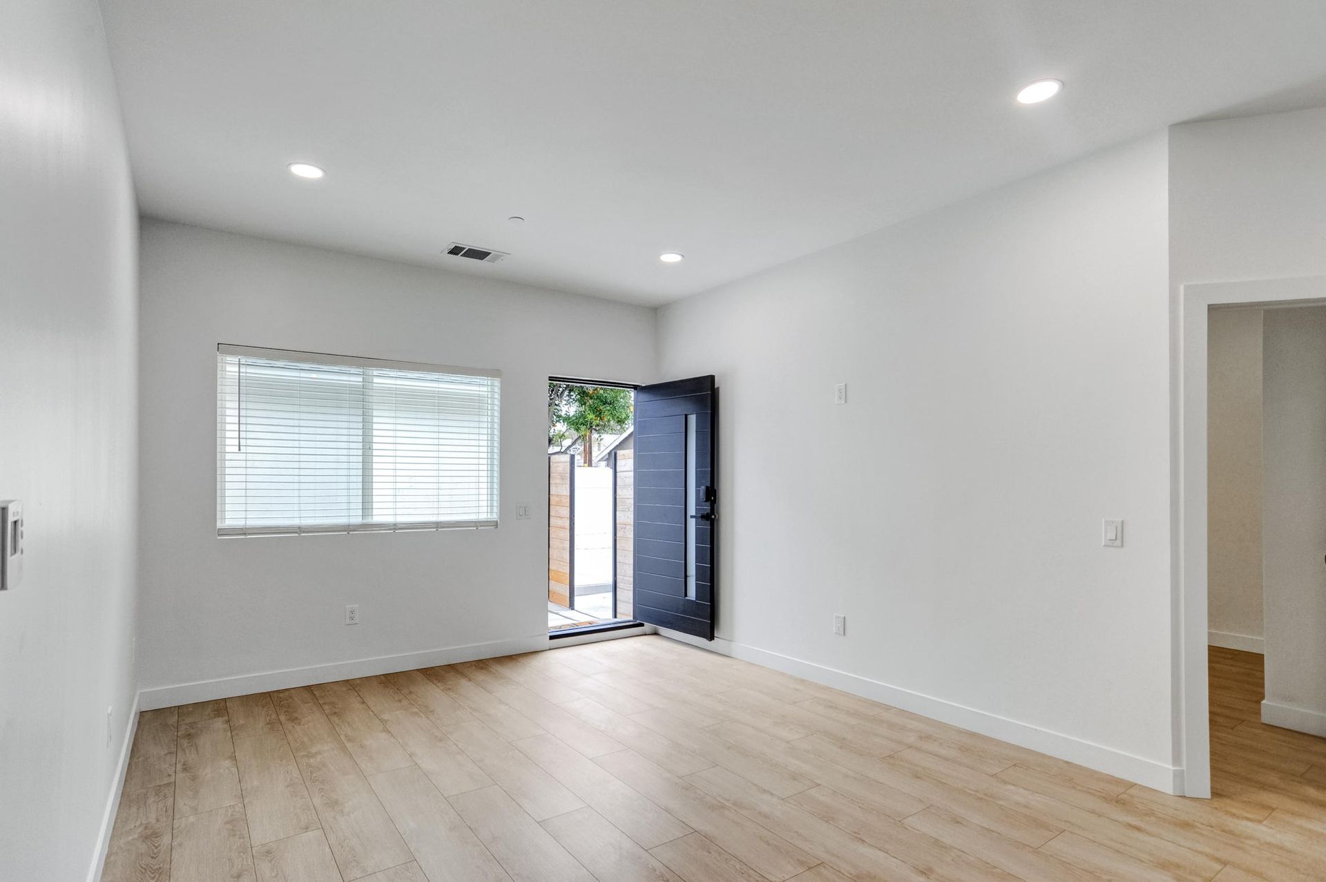 Bright empty living room with wood floors, white walls, window blinds, and open front door.