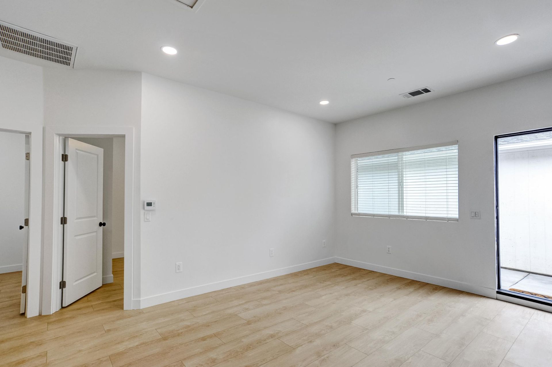 Empty bright living room with white walls, light wood floor, recessed lights, window, and sliding glass door.