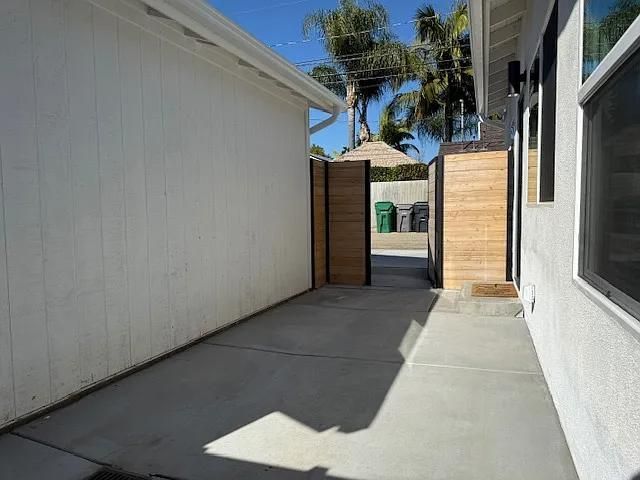 Narrow concrete walkway between white buildings leading to a wooden gate, with palm trees and blue sky beyond.