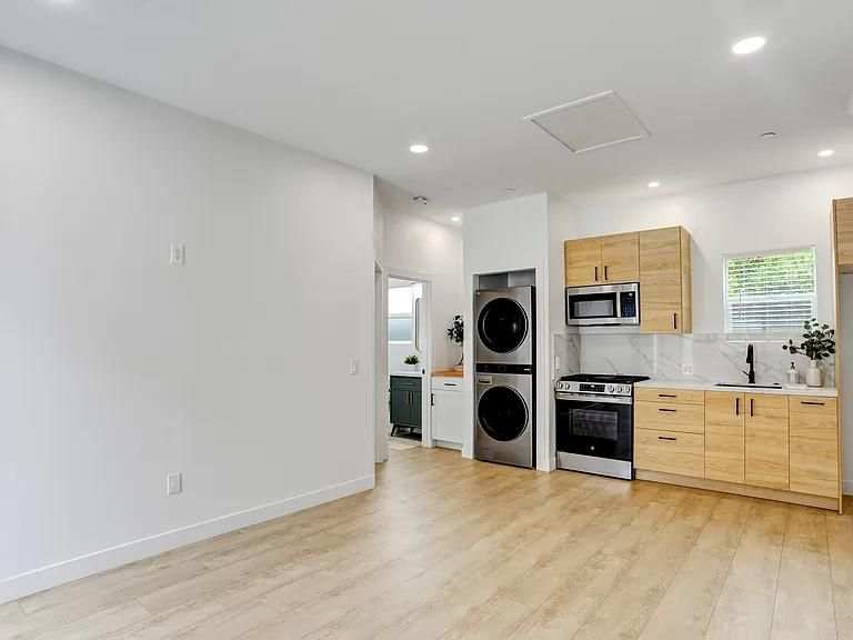 Empty modern kitchen-living space with light wood cabinets, stacked washer-dryer, and large white walls