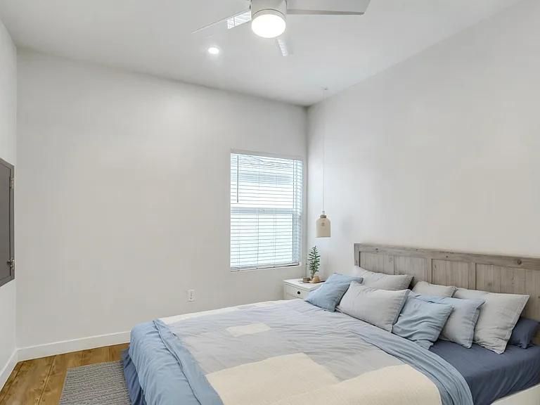 Bright bedroom with a bed, white walls, wood floor, and a window with blinds