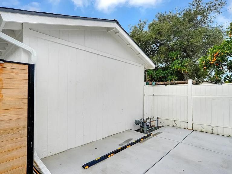 Empty concrete patio beside a white shed and fence, with a tree visible in the background.