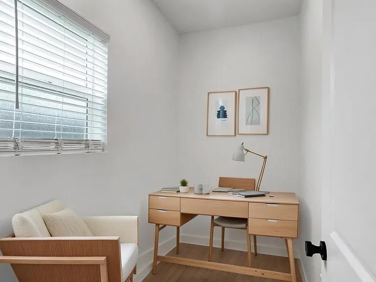 Minimal home office with wooden desk, chair, armchair, wall art, and large window with blinds.