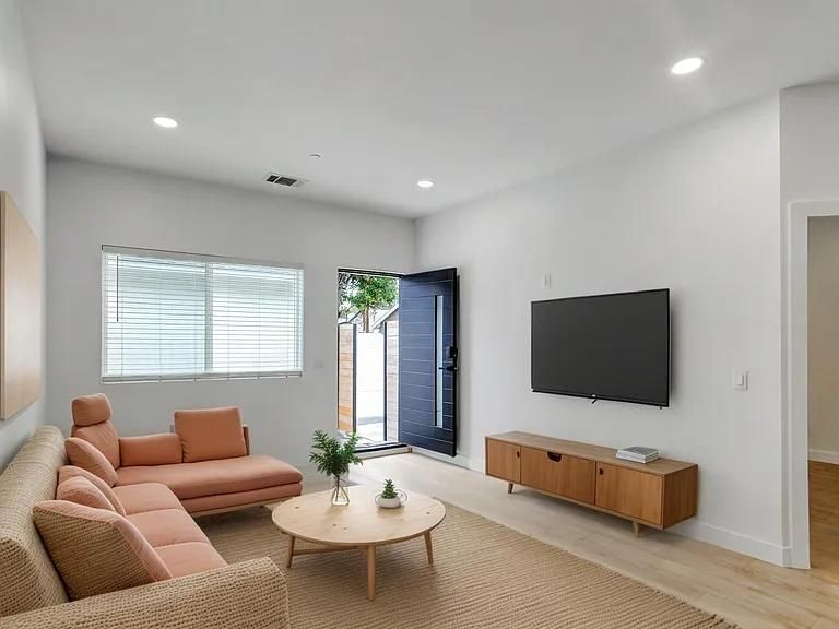 Bright living room with beige sofa, round coffee table, wall-mounted TV, and open blue front door.