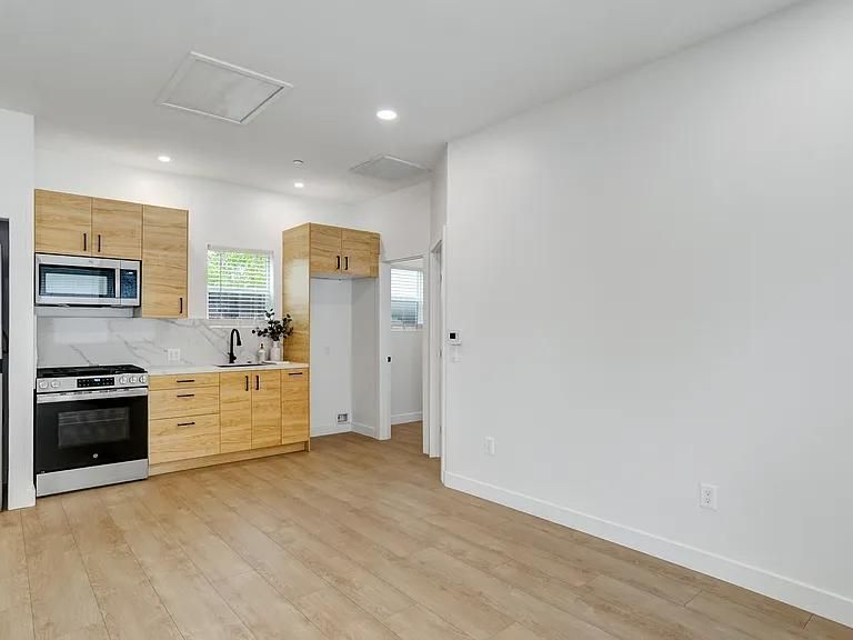 Empty modern kitchen and living area with light wood floors, white walls, and stainless steel appliances
