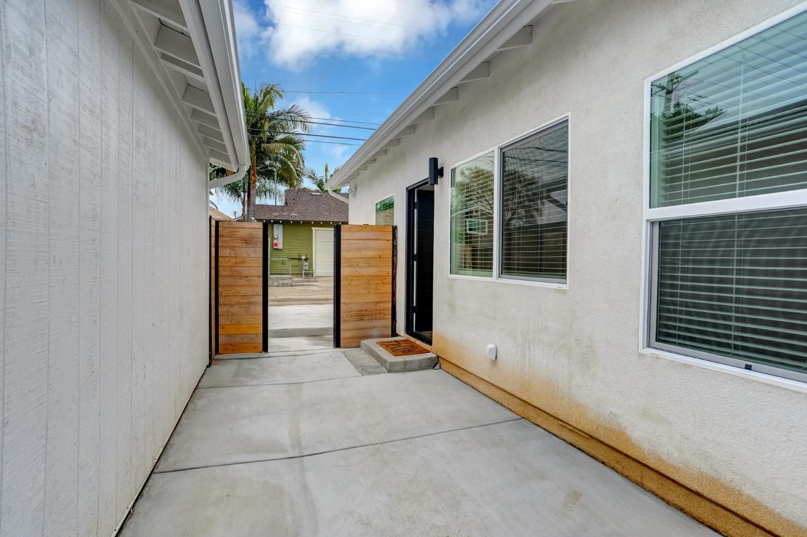 Narrow concrete walkway between white buildings leading to a wooden gate and small patio.