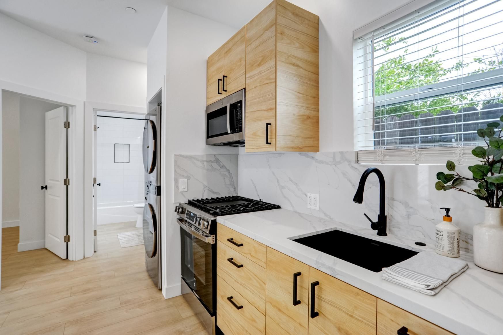 Modern kitchen with light wood cabinets, white countertops, black fixtures, stainless appliances, and a window.