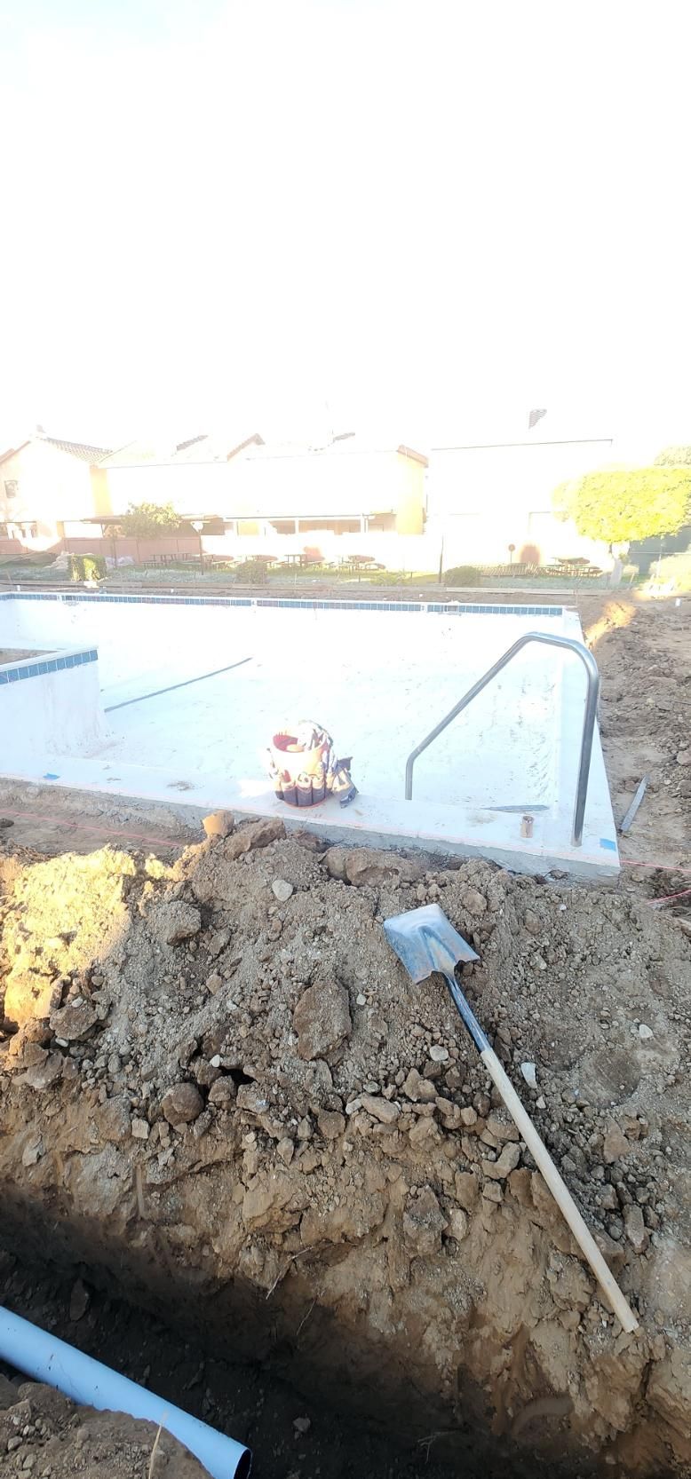 Outdoor pool under construction with muddy soil, a metal ladder, and a shovel in the foreground