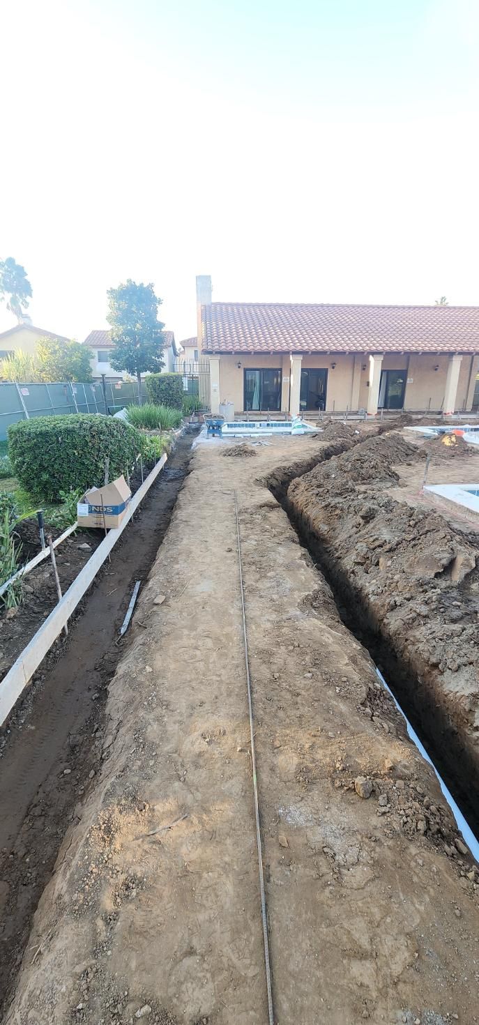 Dirt path leading to a small house with a tiled roof, flanked by garden beds and a hedge