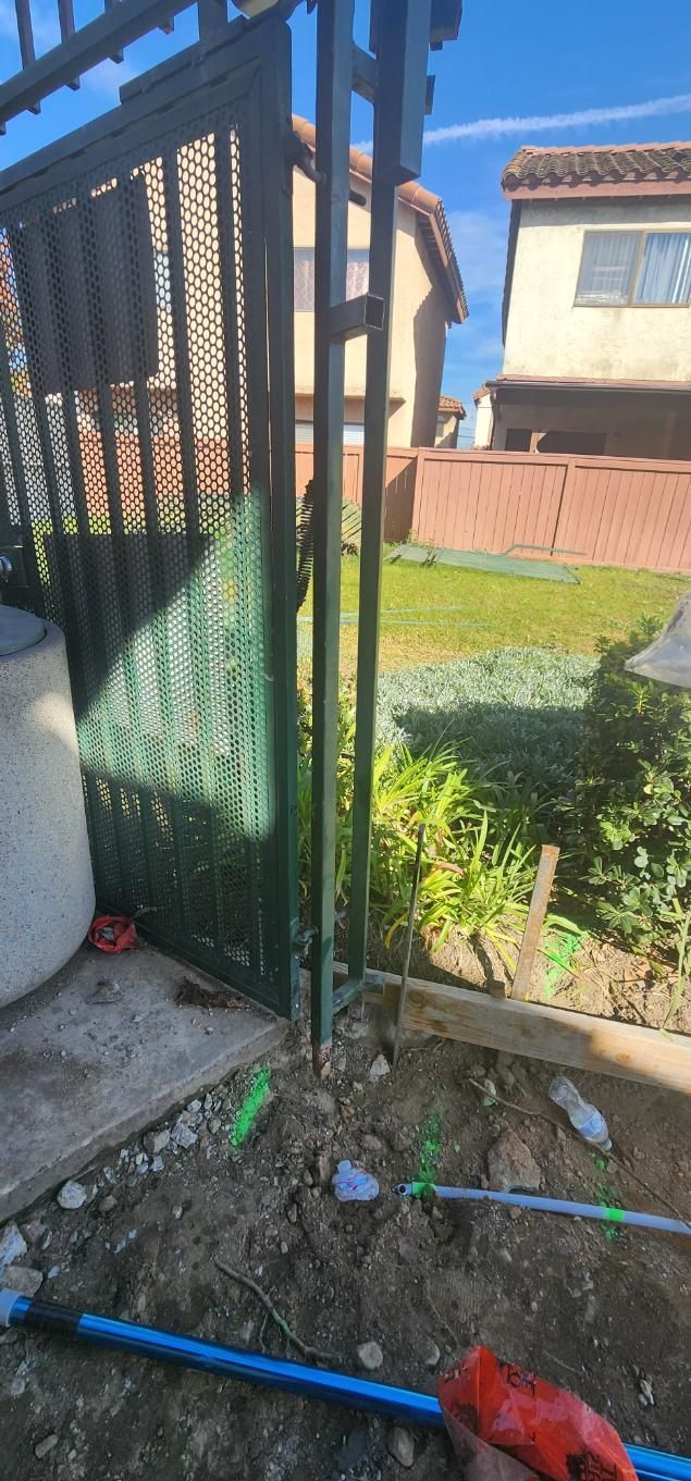 Open green metal gate beside a yard with grass, brick wall, and blue hose on the ground.