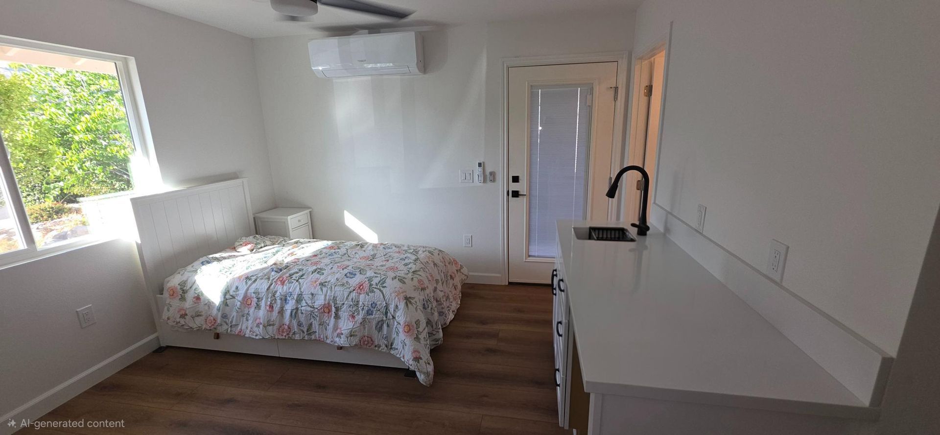 Bright bedroom with a bed, desk, window, and door, featuring white walls and wood flooring