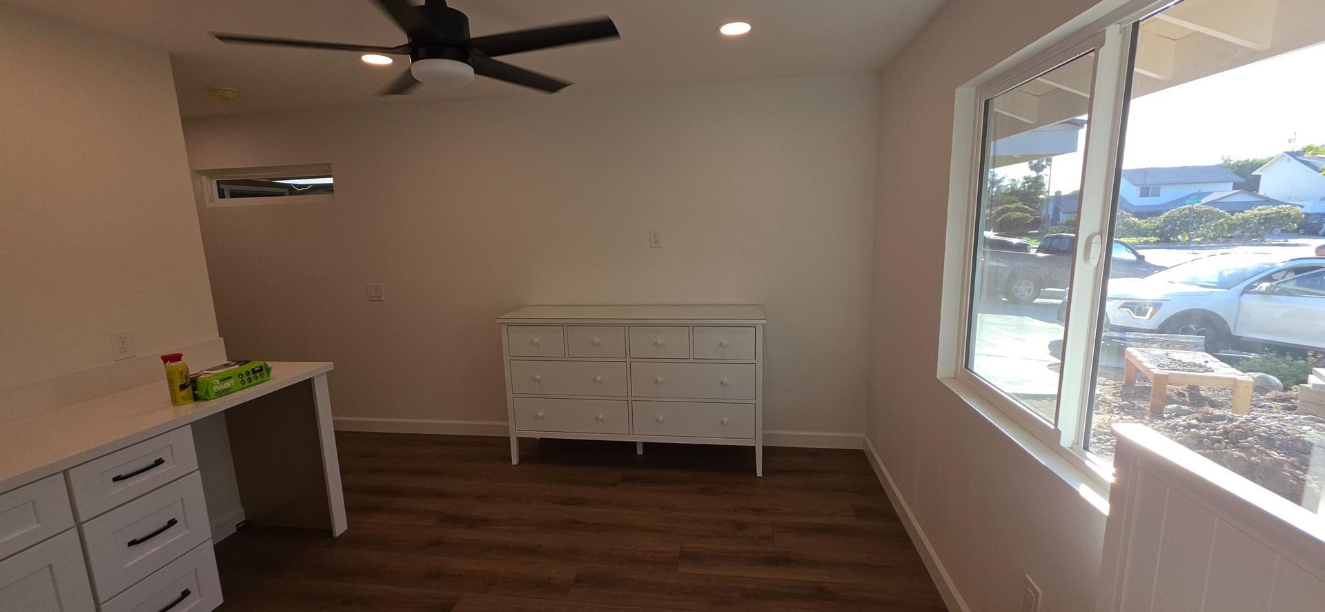 Empty modern room with wood floor, ceiling fan, white furniture, and large windows overlooking water.